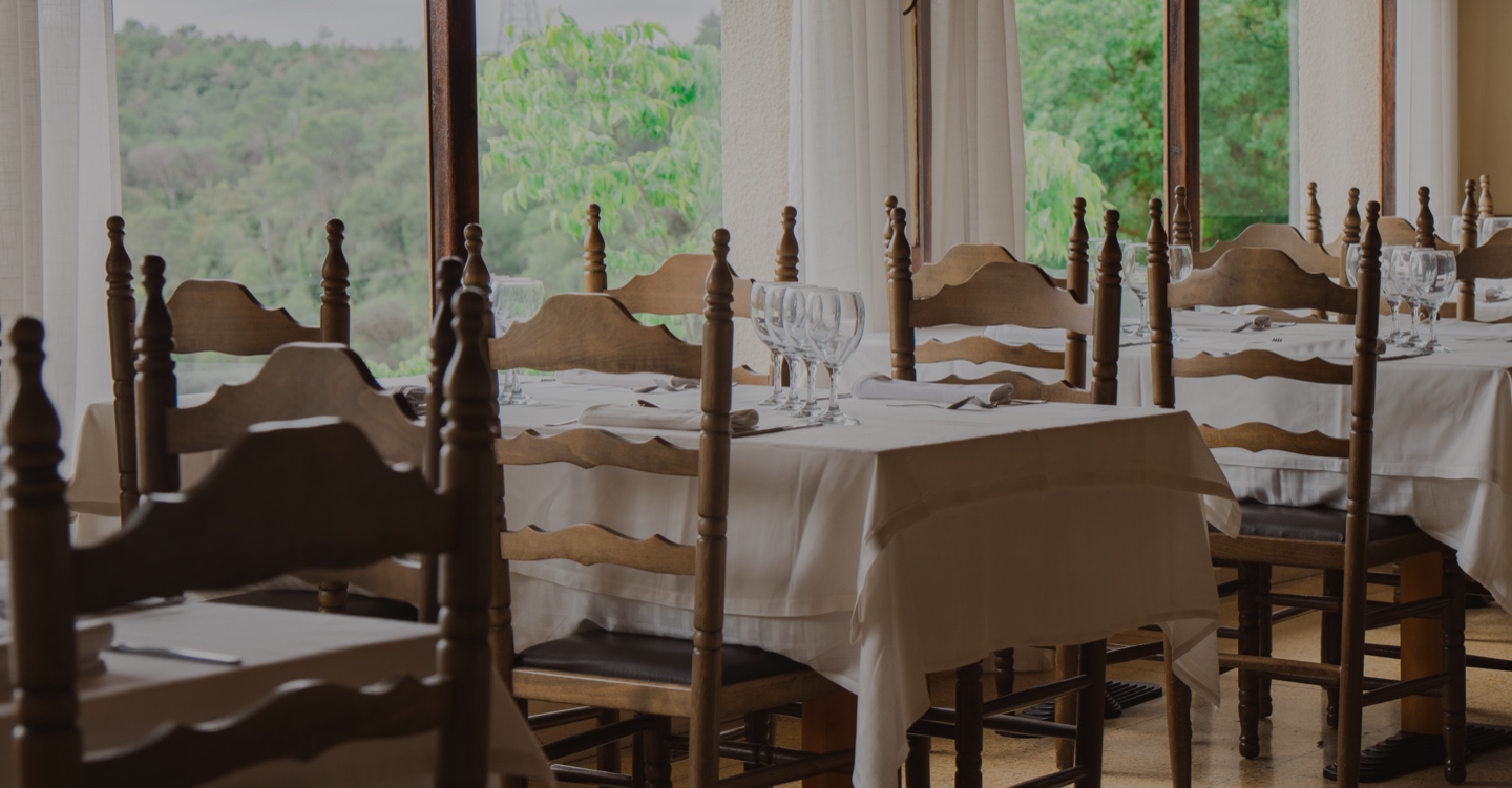 Una sala d'un restaurant amb taules i cadires de fusta tradicionals, ambientada en un entorn rural. Les finestres ofereixen una vista panoràmica d'un paisatge verd i boscós.